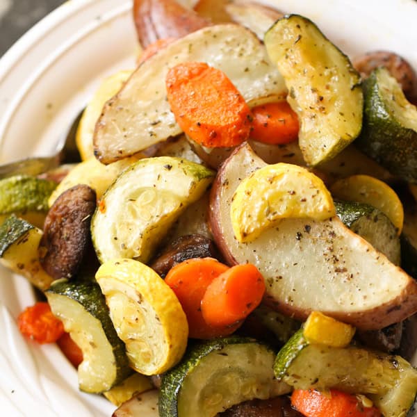 A white bowl filled with roasted vegetables, including potatoes, zucchini, carrots, and yellow squash, seasoned with spices.