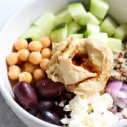 A bowl with hummus, olives, chickpeas, cucumber, quinoa, red onion, and feta cheese.