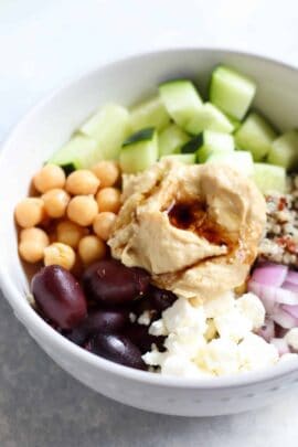 A bowl with hummus, olives, chickpeas, cucumber, quinoa, red onion, and feta cheese.