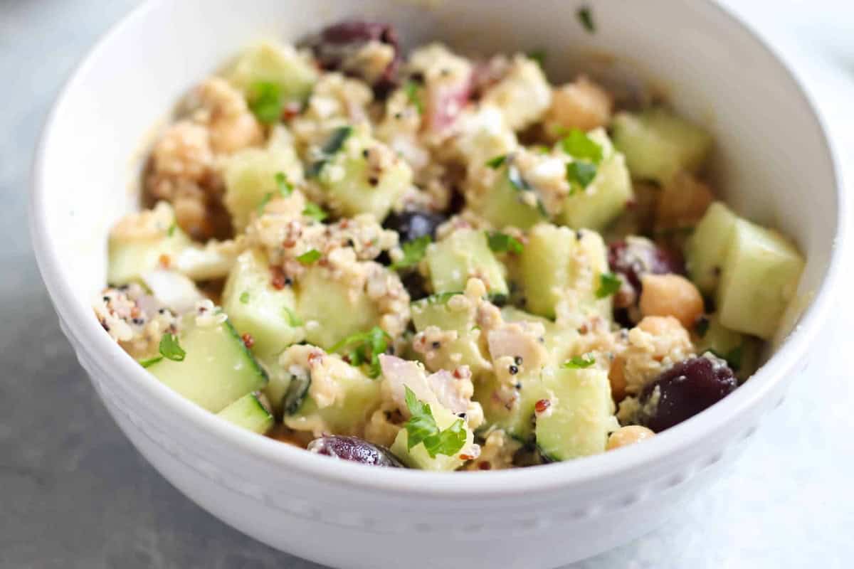 A white bowl filled with a quinoa salad containing cucumbers, chickpeas, herbs, and sliced olives.