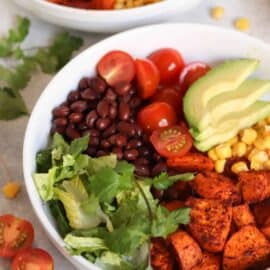 A bowl of salad with sliced avocado, corn, cherry tomatoes, black beans, cilantro, lettuce, and seasoned roasted sweet potatoes.