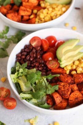 A bowl of salad with sliced avocado, corn, cherry tomatoes, black beans, cilantro, lettuce, and seasoned roasted sweet potatoes.