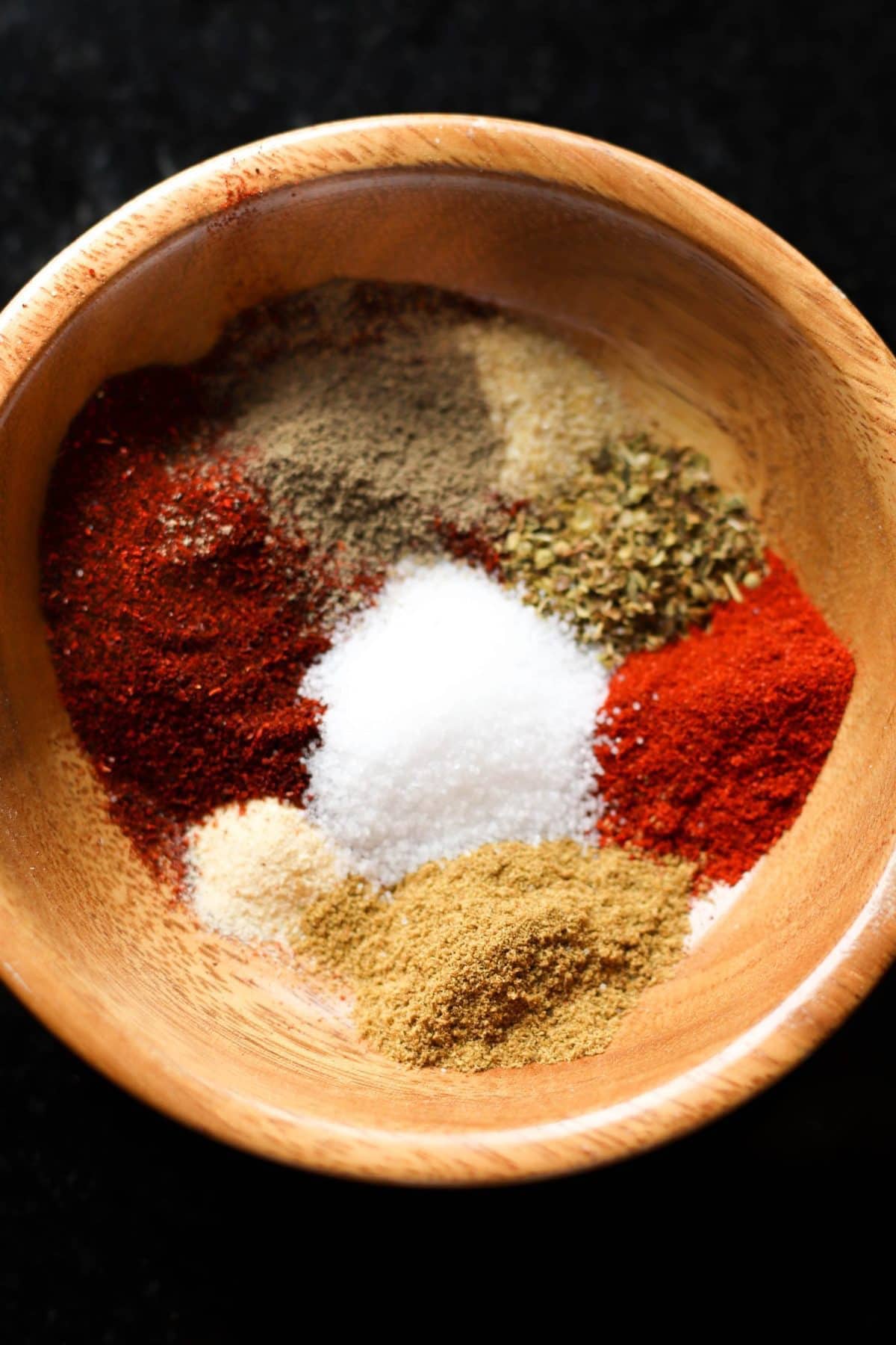 A wooden bowl filled with various spices, including salt, red chili powder, black pepper, oregano, and cumin.