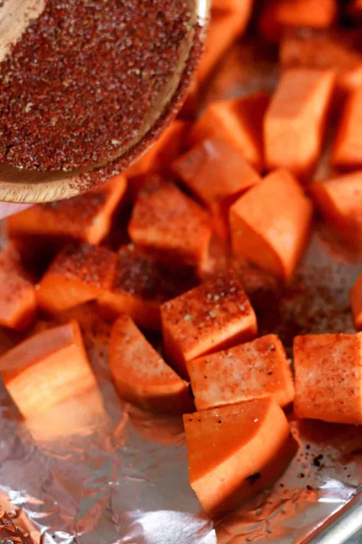 Close-up of cubed sweet potatoes on foil, being sprinkled with a red spice mix from a wooden spoon.