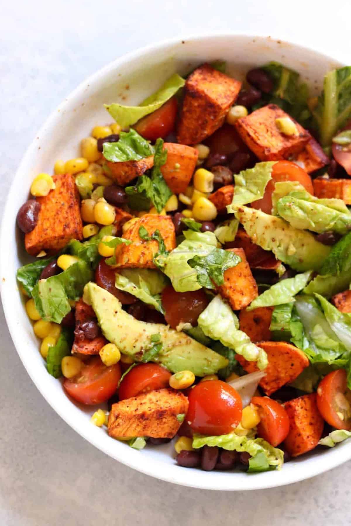 A bowl of salad with lettuce, cherry tomatoes, avocado slices, roasted sweet potato chunks, corn, and black beans.