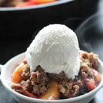 A white bowl filled with a fruit crumble dessert topped with a scoop of vanilla ice cream. Steam is visible rising from the dish.