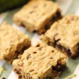 Four square-shaped zucchini chocolate chip bars placed on a striped cloth with shredded zucchini and chocolate chips scattered around.