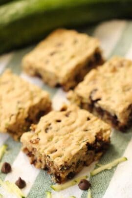 Four square-shaped zucchini chocolate chip bars placed on a striped cloth with shredded zucchini and chocolate chips scattered around.