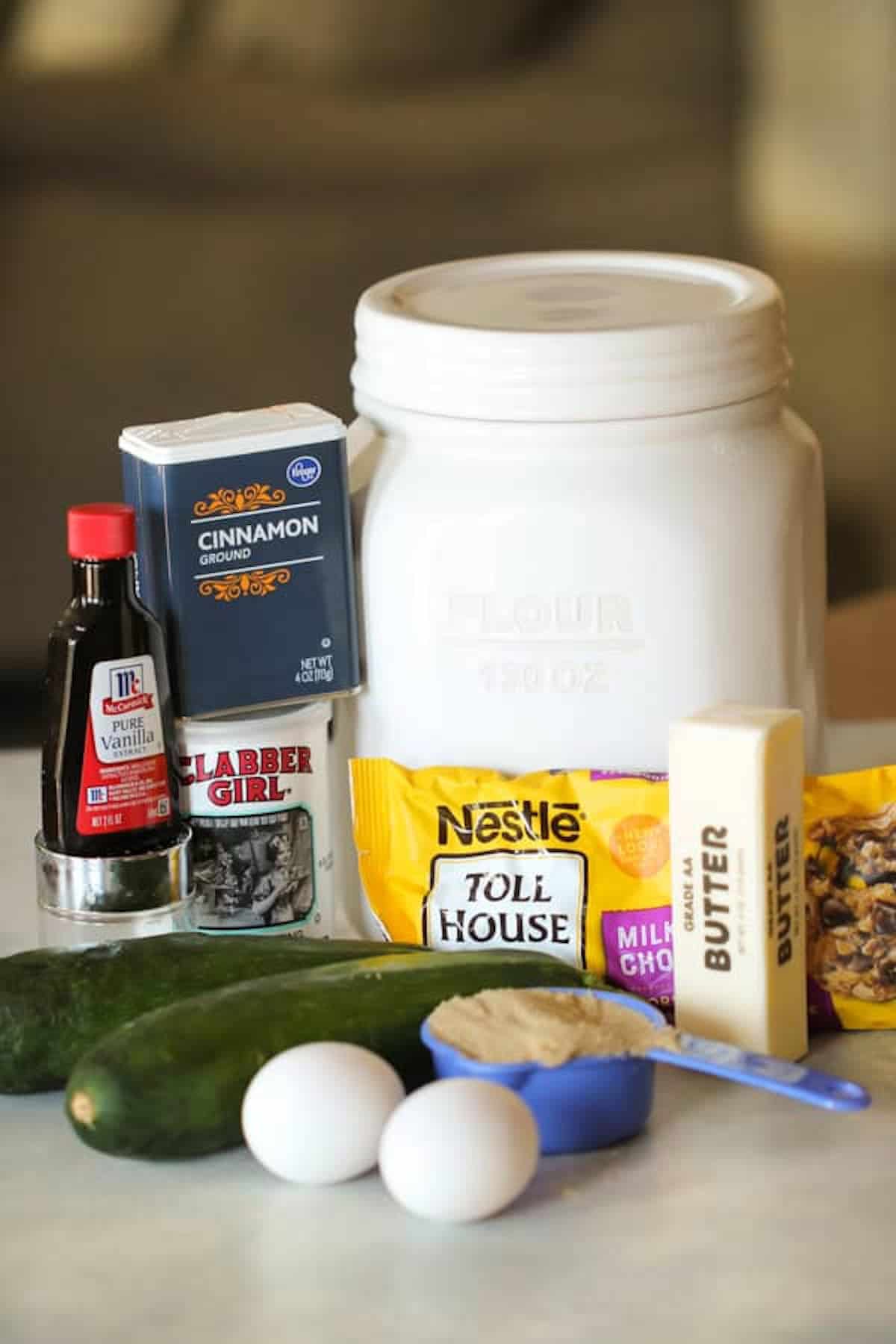 A collection of baking ingredients on a countertop, including flour, ground cinnamon, pure vanilla extract, baking powder, eggs, brown sugar, butter, a zucchini, and chocolate chips.