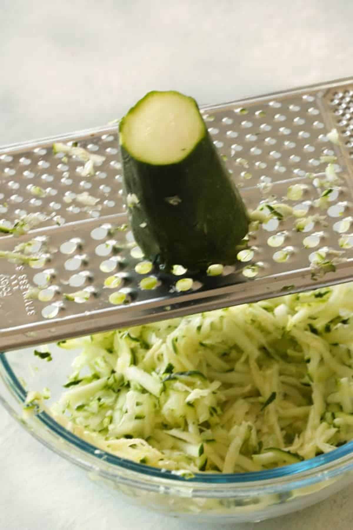 A partially grated zucchini on a metal grater positioned over a glass bowl filled with shredded zucchini.