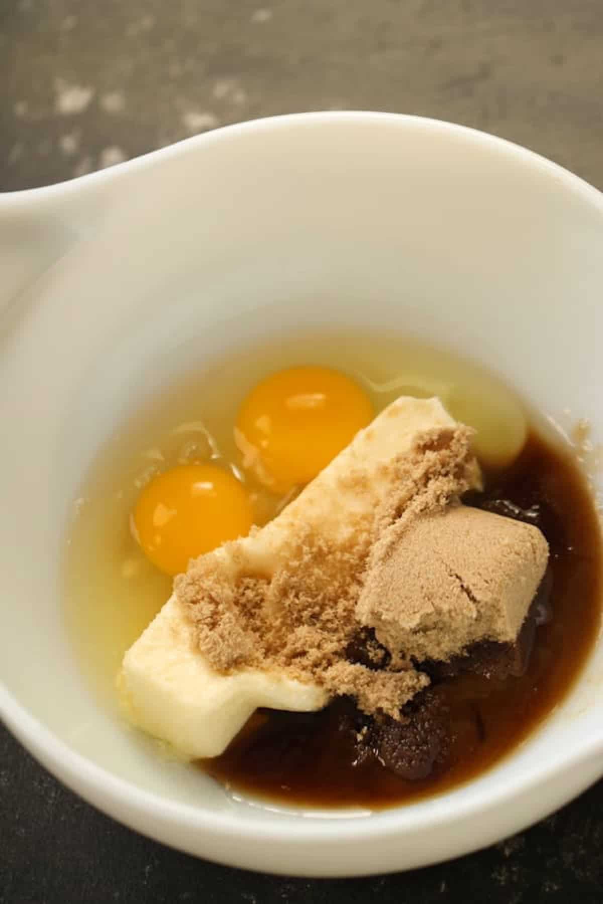 A white mixing bowl contains two raw eggs, a stick of butter, brown sugar, and a liquid ingredient.