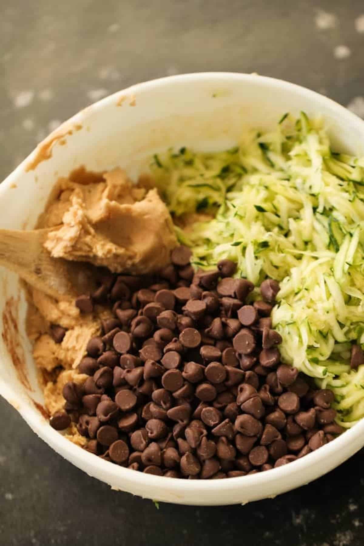 A mixing bowl contains peanut butter, shredded zucchini, and chocolate chips with a wooden spoon.