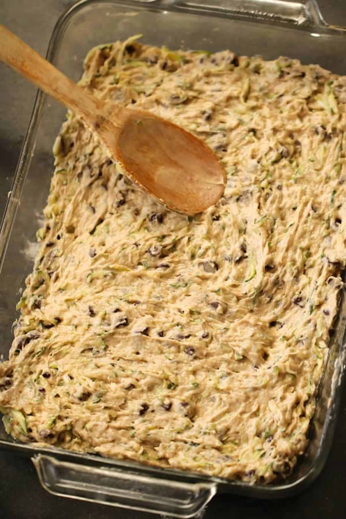 A glass baking dish filled with a mixture of batter and chocolate chips, being leveled with a wooden spoon.