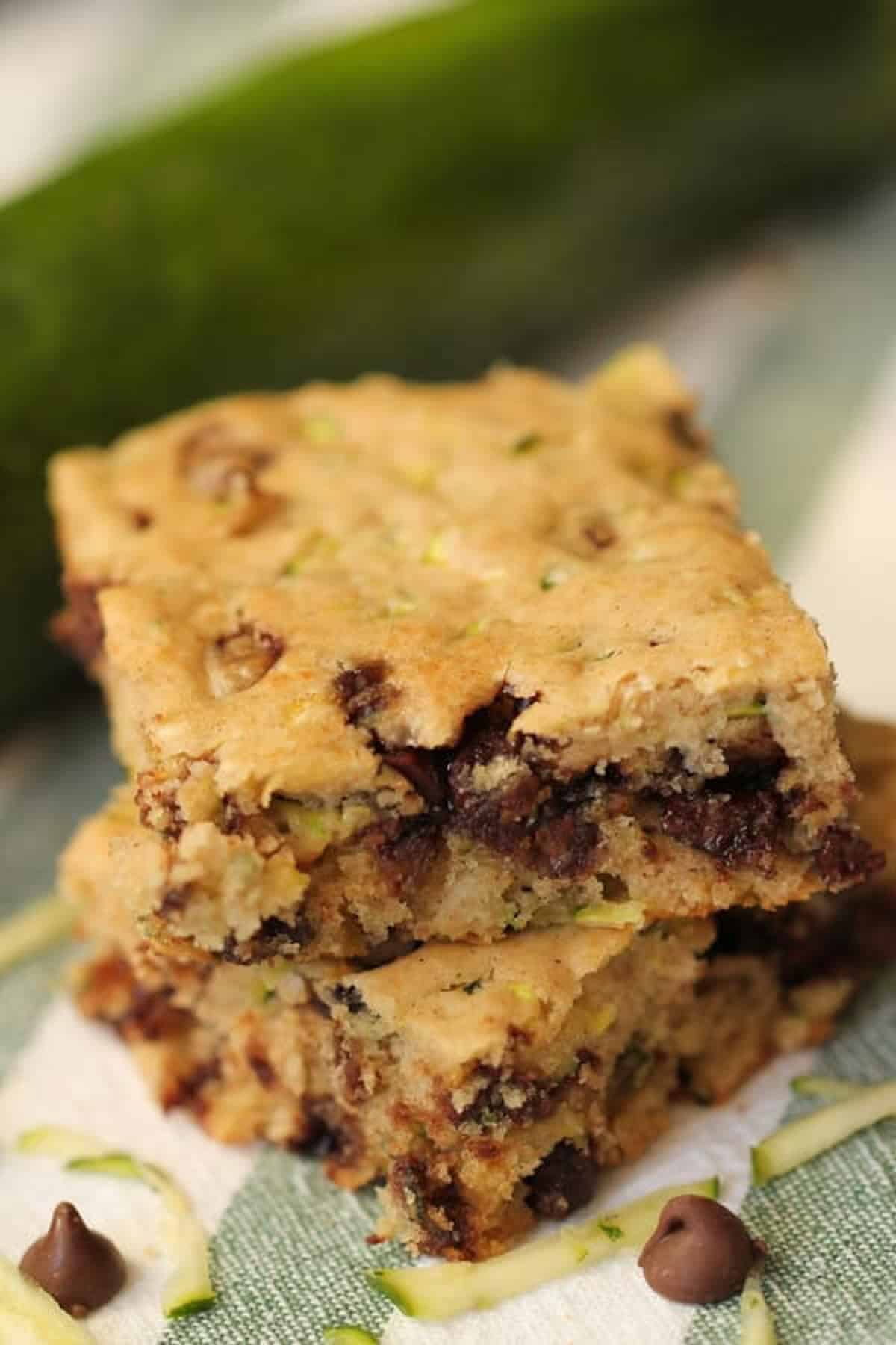 Two stacked zucchini chocolate chip bars on a white and green cloth, with a whole zucchini in the background.