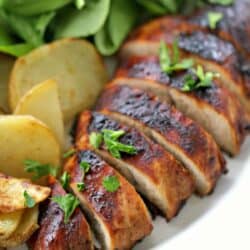 Sliced, marinated turkey breast garnished with herbs, served with roasted potato slices and a side of fresh spinach leaves on a white plate.