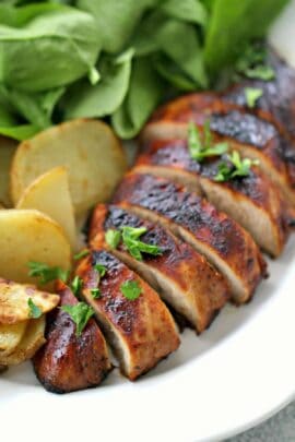 Sliced, marinated turkey breast garnished with herbs, served with roasted potato slices and a side of fresh spinach leaves on a white plate.