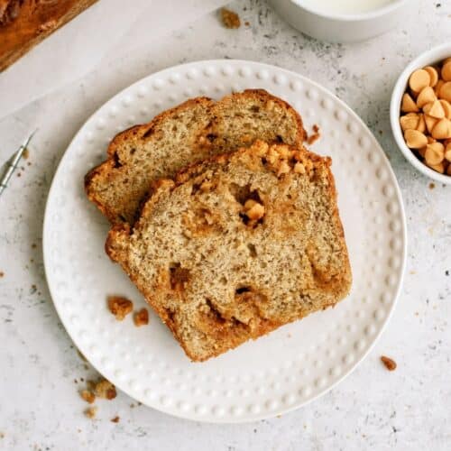 Two slices of Banana Bread on a small white plate with a cup of milk and a small cup of butterscotch chips on the side.