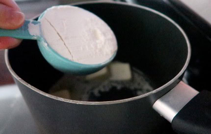 Person adding a blue cup of flour into a black pot with melted butter on a stove.