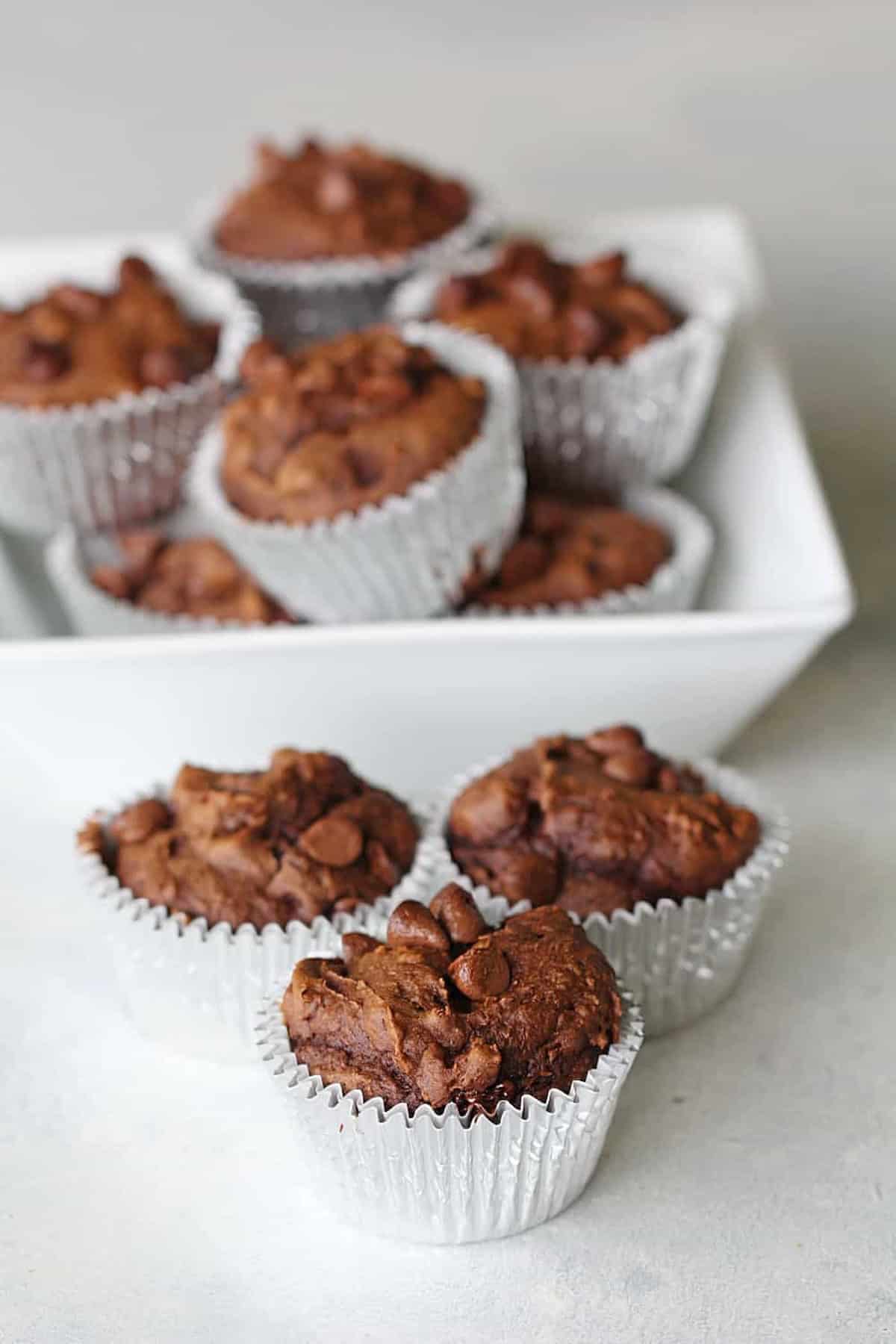 A white dish holding multiple chocolate muffins in paper liners with three additional muffins in the foreground on a light surface.