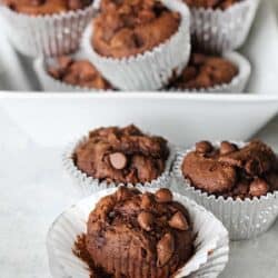 A white tray with multiple chocolate muffins in silver foil liners, with three muffins in the foreground and several more stacked in the background.