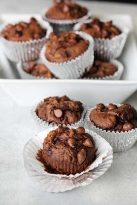 A white tray with multiple chocolate muffins in silver foil liners, with three muffins in the foreground and several more stacked in the background.