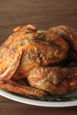 A whole roasted chicken with crispy skin, garnished with herbs, placed on a white plate.