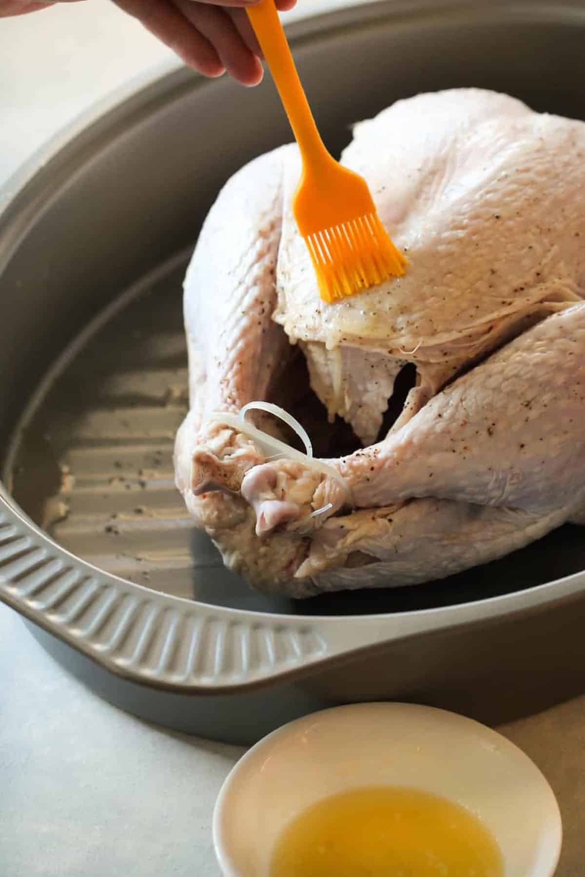 A raw turkey in a roasting pan is being brushed with butter. A small bowl with melted butter is nearby.