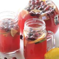 Glass pitcher and two mason jars filled with red cranberry drink, garnished with lime and lemon slices. Cranberries are visible in the drink. Whole lime and lemon are nearby.