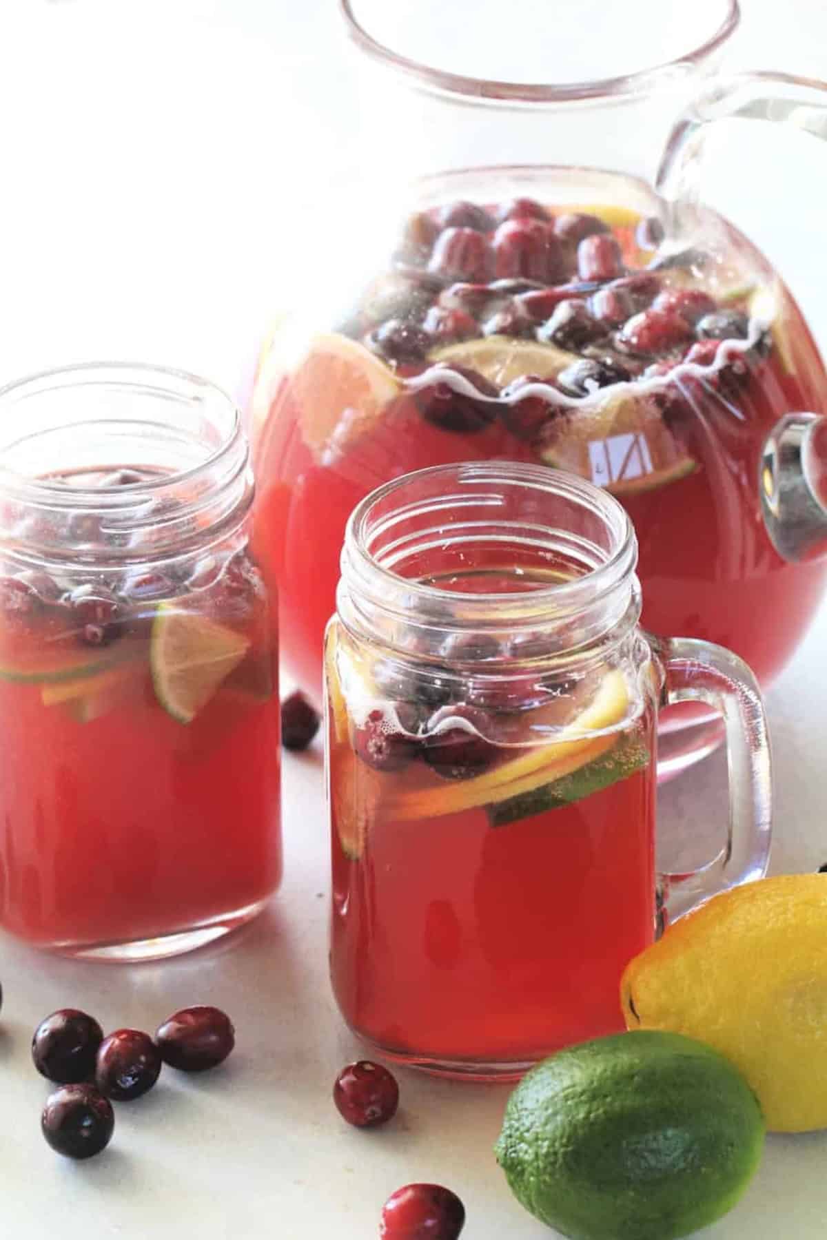 Glass pitcher and two mason jars filled with red cranberry drink, garnished with lime and lemon slices. Cranberries are visible in the drink. Whole lime and lemon are nearby.