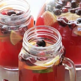 Glass pitcher and two mason jar mugs filled with cranberry beverage, garnished with lemon and lime slices, and cranberries.