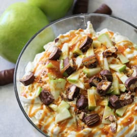 A bowl of caramel apple salad garnished with chopped pieces of apples and chocolate bars. Two green apples and pieces of chocolate bars are placed next to the bowl.