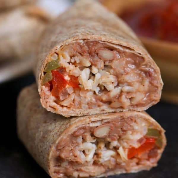 A close-up of a halved burrito showing a filling of beans, rice, cheese, and diced vegetables. A bowl of salsa is blurred in the background.