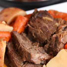 A plate of pot roast with chunks of beef, carrots, and potatoes in front of an electric pressure cooker.