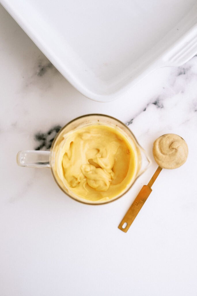 A clear measuring cup filled with creamy yellow mixture next to a teaspoon of the same mixture, placed on a marble surface.