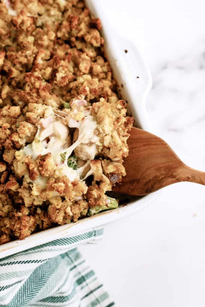 A wooden spoon is serving a portion of casserole with a breadcrumb topping from a white baking dish. The dish is placed on a white surface with a green-striped cloth nearby.