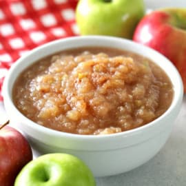 A bowl of applesauce is surrounded by whole red and green apples, with a red and white checkered cloth in the background.