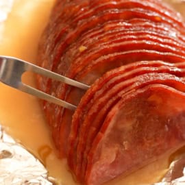 A metal fork holds a sliced ham that is arranged on a piece of foil, covered in a glossy glaze, with juices pooling around it.