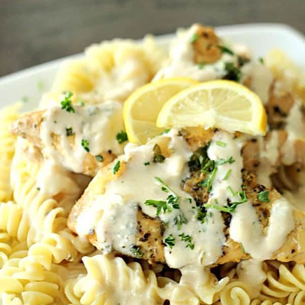 A plate of pasta topped with grilled chicken, creamy white sauce, chopped parsley, and lemon slices.