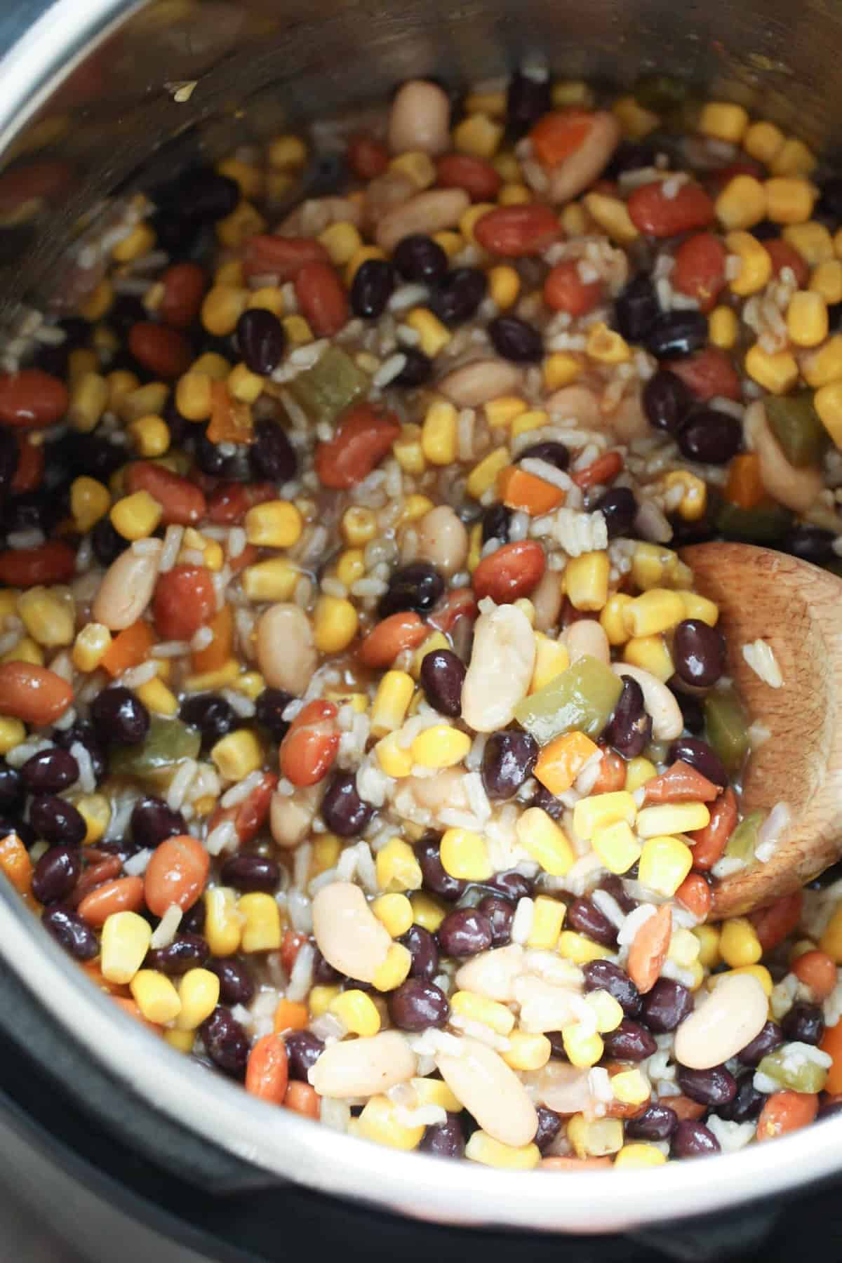 A pot filled with mixed beans, corn, and green peppers being stirred with a wooden spoon.