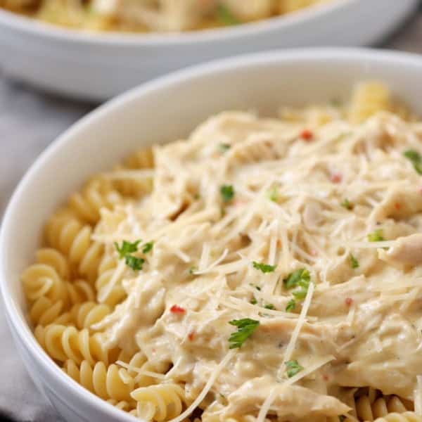 A white bowl filled with rotini pasta topped with a creamy sauce and garnished with green herbs. Another similar bowl is partially visible in the background.