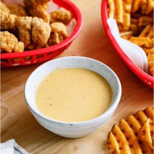 A bowl of Copycat Chick-Fil-A Sauce surrounded by a basket of chicken tenders and a basket of french fries.