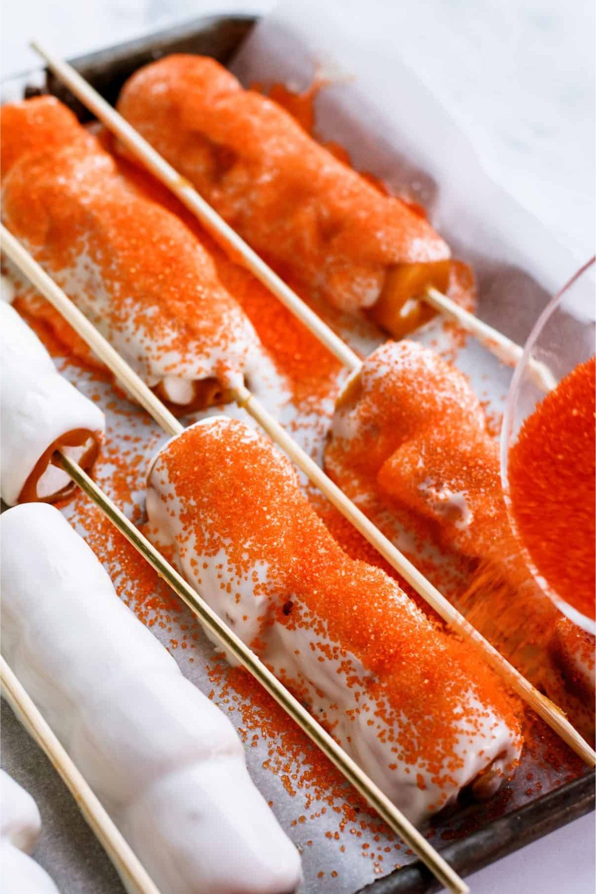 Sprinkling orange sprinkles from a small glass bowl on top of white chocolate covered skewers.