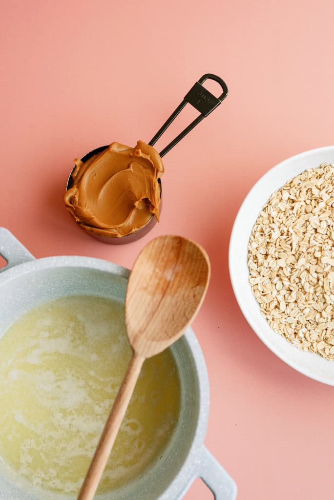 Butter melted in a saucepan with a wooden spoon sitting on top. A measuring cup with peanut butter and a small bowl of quick oats.