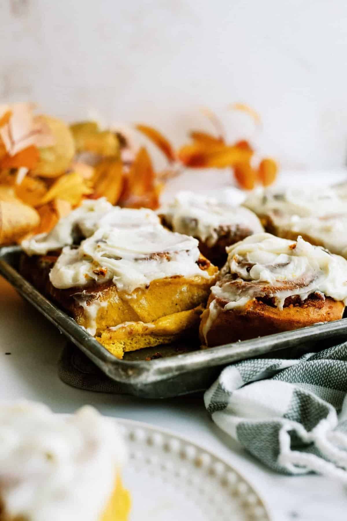 A tray of freshly baked cinnamon rolls with thick white icing, set on a table adorned with autumn leaves. A partly eaten cinnamon roll is in focus, with a plate and cloth napkin nearby.