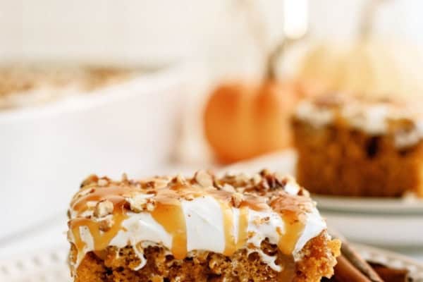 A slice of pumpkin cake topped with frosting and a drizzle of caramel sauce on a white plate, with more cake and a pumpkin in the blurred background.