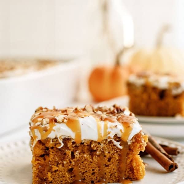 A slice of pumpkin cake topped with frosting and a drizzle of caramel sauce on a white plate, with more cake and a pumpkin in the blurred background.