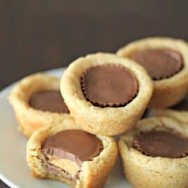 Plate of five peanut butter cup cookies, one with a bite taken out. Cookies have soft, golden-brown edges with whole peanut butter cups in the center.