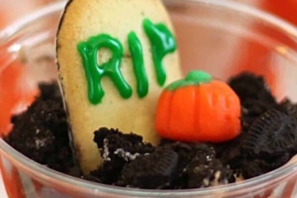 A dessert cup resembling a gravesite, with a cookie "tombstone" labeled "RIP" and a candy pumpkin on top of crumbled chocolate cookies.