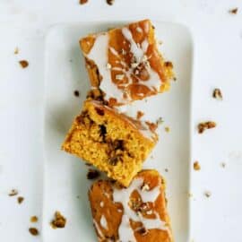 Three pieces of glazed pumpkin cake with nuts, arranged on a white rectangular plate. Crumbs and nuts scattered around.