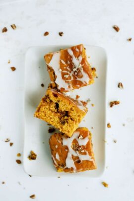 Three pieces of glazed pumpkin cake with nuts, arranged on a white rectangular plate. Crumbs and nuts scattered around.